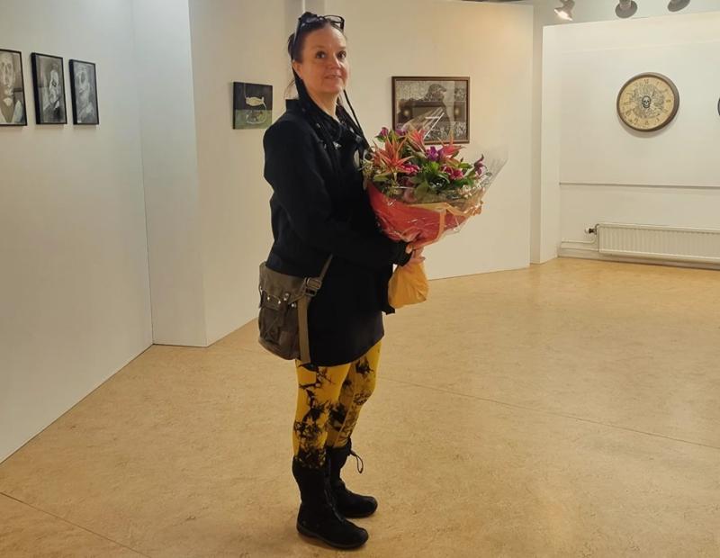 En kvinna står i ett konstgalleri med en blombukett i handen, klädd i en mörk kappa, gulblommiga leggings och svarta stövlar. På galleriets väggar hänger tavlor och klockor.
