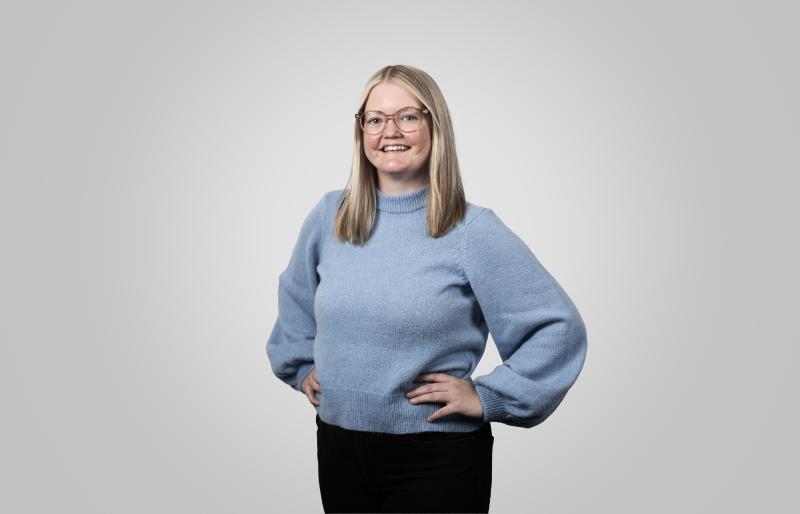 En person med rakt, axellångt blont hår och glasögon står och ler, klädd i en ljusblå tröja och svarta byxor mot en enfärgad, ljusgrå bakgrund.