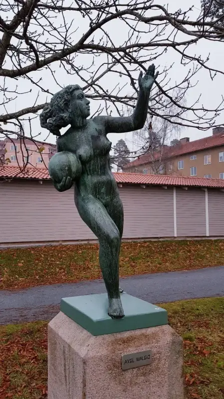 En bronsstaty av en kvinna som håller en boll, stående på en sockel utomhus med byggnader, ett träd och en stig i bakgrunden.