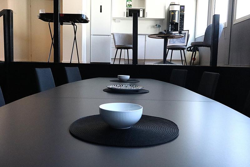 En modern matplats med ett långt bord, svarta bordsunderlägg och vita skålar, med ett litet kök och ett runt bord i bakgrunden.
