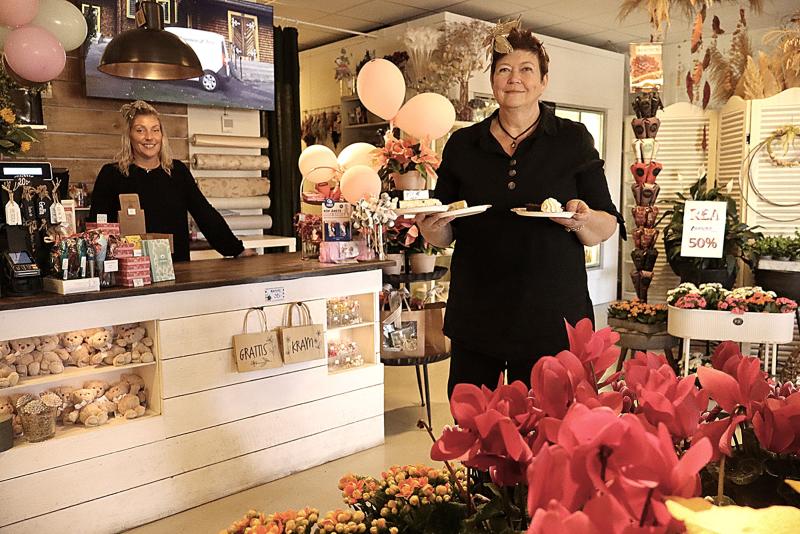 Två kvinnor i en blomsteraffär; den ena står bakom disken, den andra håller i tallrikar med tårta. Butiken är dekorerad med blommor, växter, ballonger och presentartiklar.