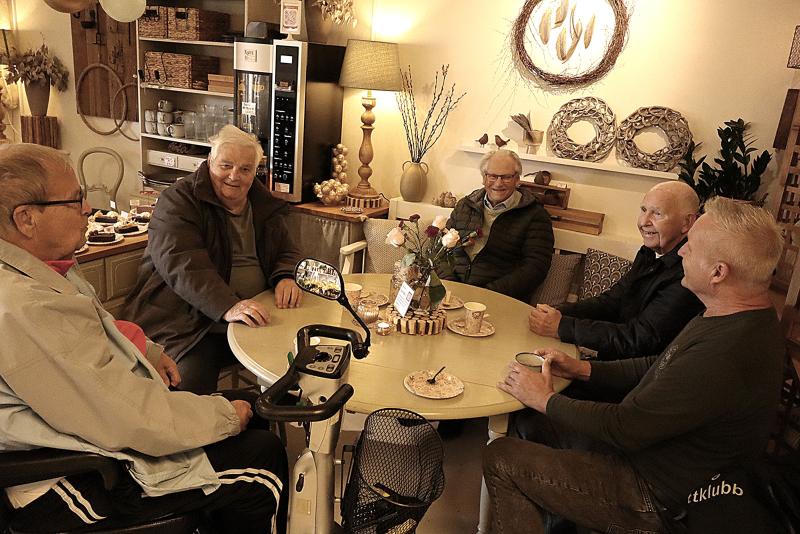 Fem äldre män sitter runt ett runt bord på ett mysigt café och dricker kaffe och samtalar. Miljön har varm belysning och dekorativa föremål på hyllor och väggar.