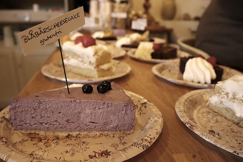 En skiva glutenfri blåbärscheesecake på en papperstallrik, märkt med en liten skylt, ligger bland olika kakor på en träbänk.