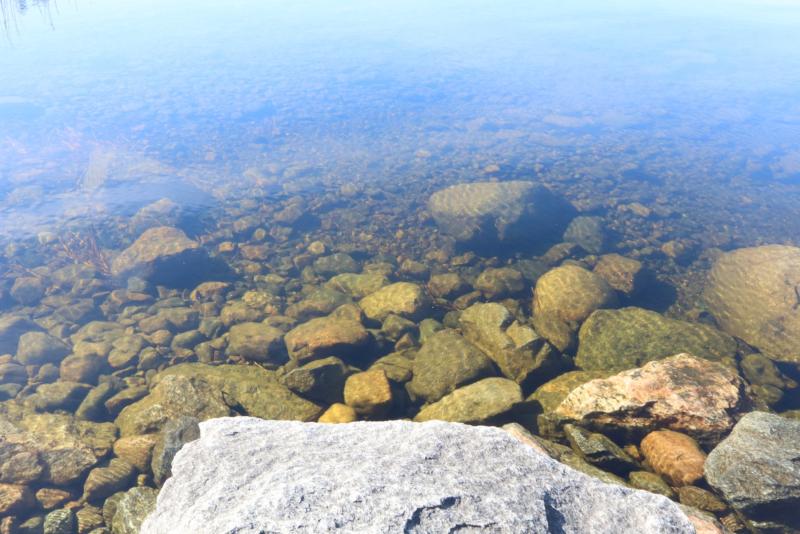 Stora stenar och många små stenar syns genom det klara vattnet vid kanten av en lugn vattensamling, där solljuset skapar mönster på de nedsänkta stenarna.