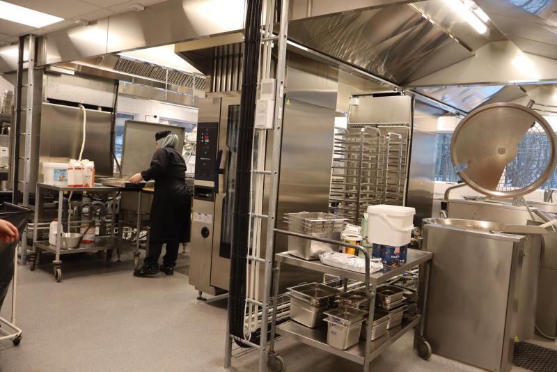 En person i ett storkök arbetar vid en bänk som omges av rostfria apparater, ställningar och olika kökstillbehör.