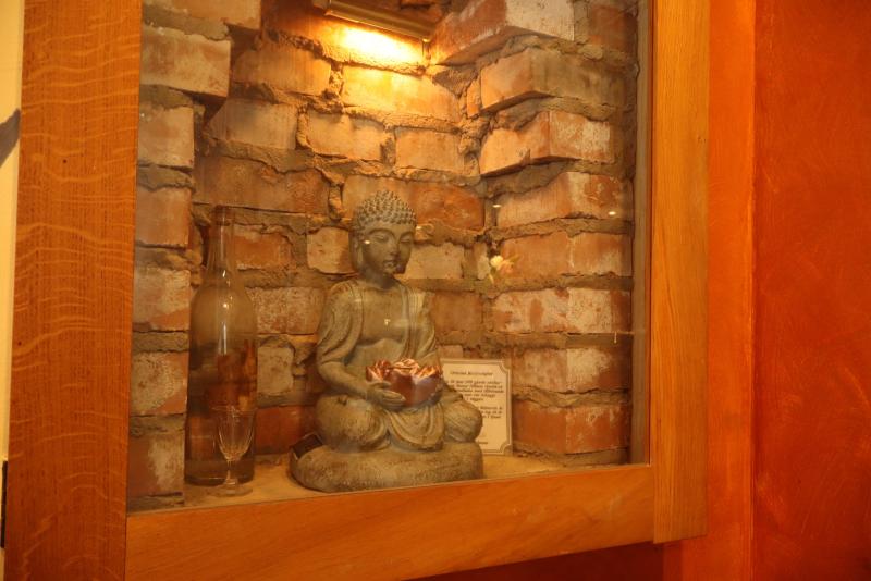 En liten Buddhastaty som håller en lotusblomma står i en glasmonter placerad i en nisch av tegel, med en flaska och en plakett bredvid sig, upplyst av ett varmt ljus.