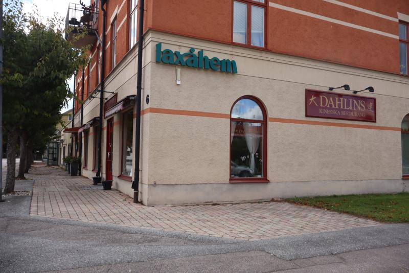 Ett gatuhörn med en tvåvåningsbyggnad i orange och beige; på skyltarna står det ”laxåhem” och ”DAHLINS KINESISKA RESTAURANG”. Man ser tegelbeläggning och grönska.