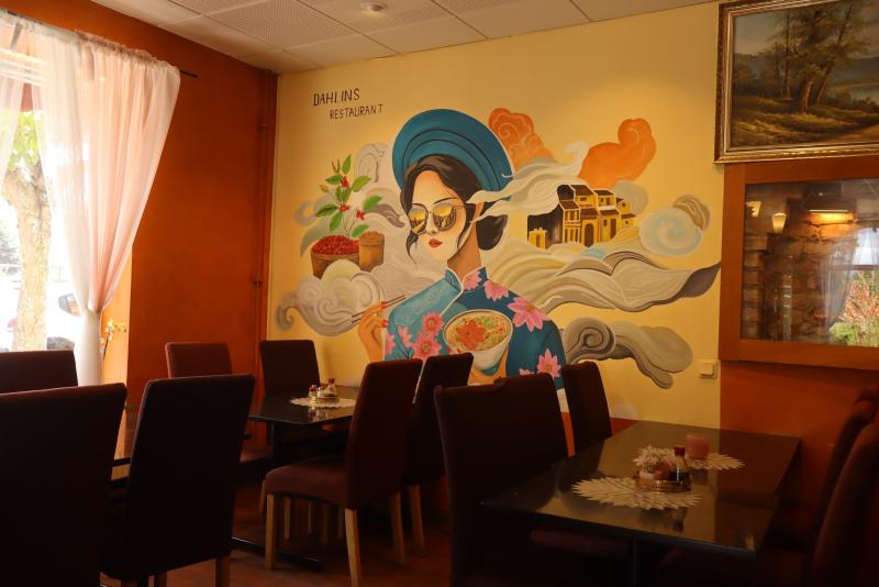 En restauranginteriör med tomma bord och stolar, där en stor väggmålning av en kvinna i traditionell dräkt syns med färgglada moln och byggnader i bakgrunden.
