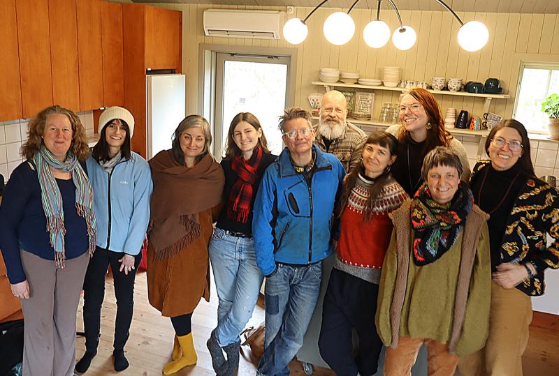 En grupp på tio vuxna poserar tillsammans i ett kök och ler mot kameran. Hyllor med disk och träskåp syns i bakgrunden.