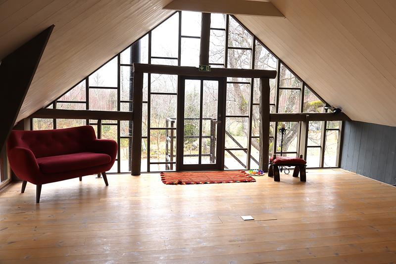 Loftrum med trägolv, en röd soffa, en liten matta, en pall, ett skrivbord och stora fönster med utsikt över träden utanför.