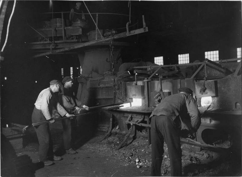 Tre arbetare använder långa verktyg för att ta hand om en stor industriugn i en svagt upplyst fabrik, med maskiner och fönster synliga i bakgrunden.