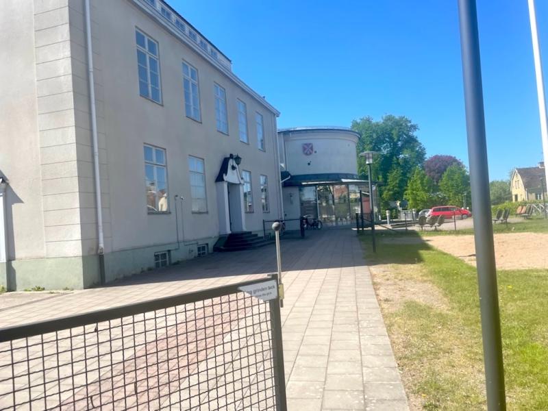 En vit byggnad i två våningar med stora fönster och en stig som leder till entrén. Scenen är klar och solig, med en gräsyta till höger och en flaggstång i förgrunden.