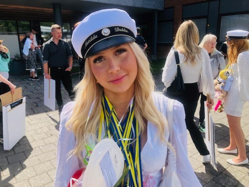 En ung kvinna med examensmössa och flera medaljer står utanför tillsammans med andra, håller i presenter och ler mot kameran.