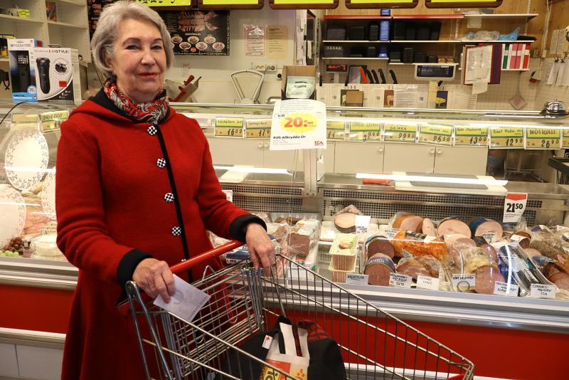 Äldre kvinna i röd rock står med en kundvagn framför en delikatessdisk med kött och ostar i en livsmedelsbutik.