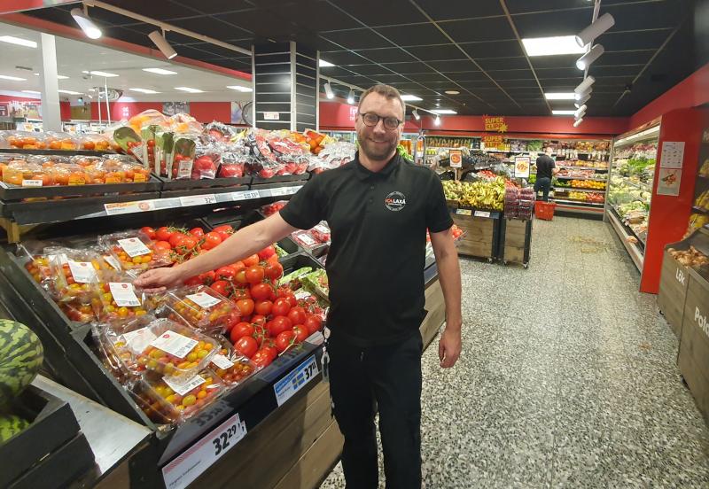 En man med glasögon och en svart polotröja står i frukt- och grönsaksavdelningen i en livsmedelsbutik och ler samtidigt som han pekar på förpackade körsbärstomater i ett skyltställ.