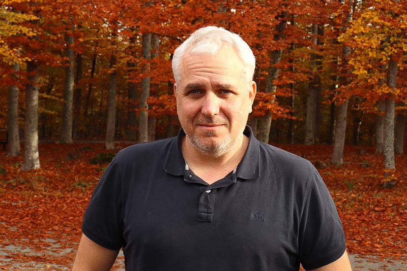 En man med kort grått hår och skägg bär en mörk polotröja och står utomhus framför träd med livfulla höstlöv.