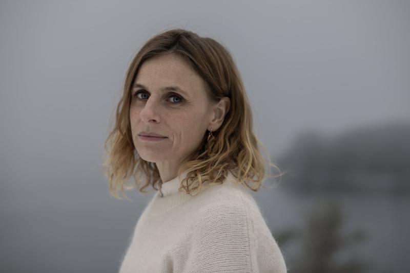 En kvinna med axellångt blont hår och en gräddfärgad tröja tittar in i kameran med ett neutralt uttryck mot en dimmig bakgrund utomhus.