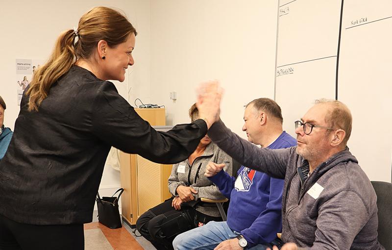 En stående kvinna ger en sittande man en high-five i ett klassrum med andra personer som sitter och tittar på.