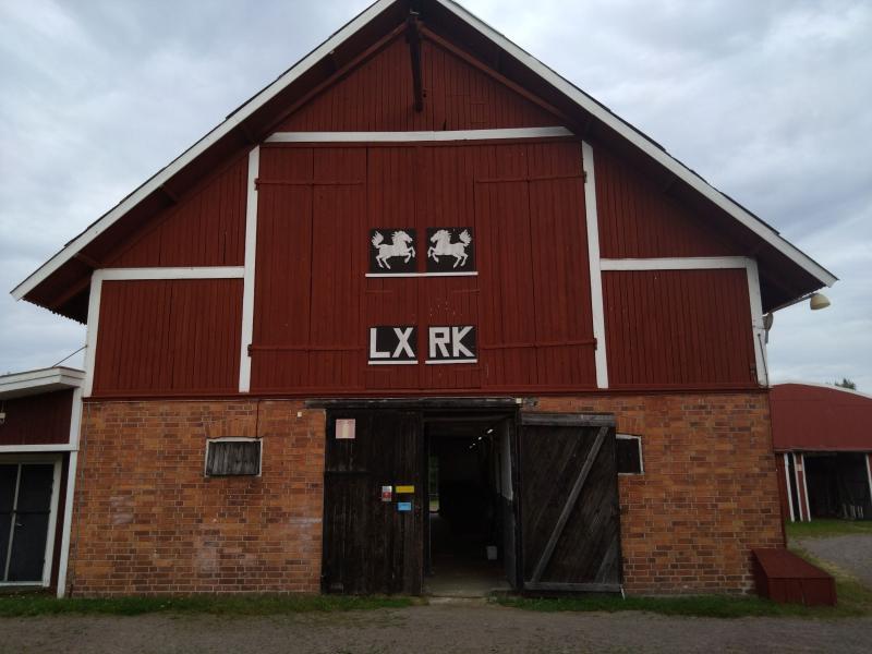 En stor röd och brun lada med två svartvita hästskyltar ovanför entrén. Ladan har delvis öppna stora trädörrar och en gräsbevuxen gång som leder till den.