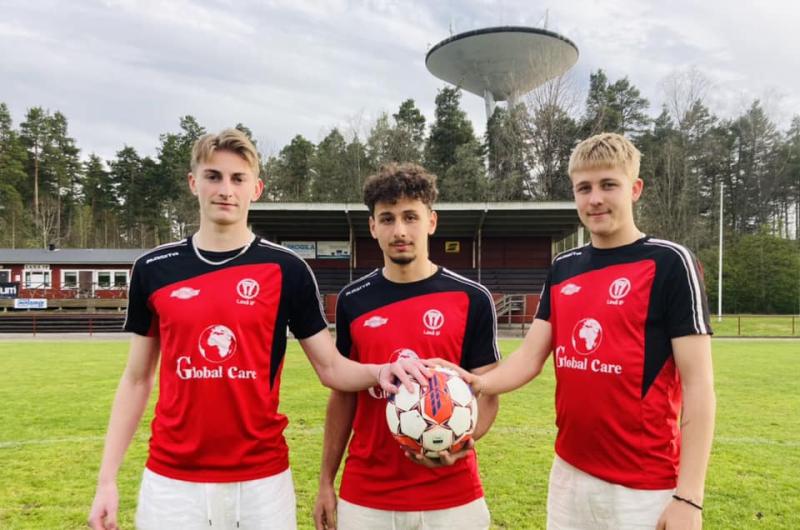 Tre unga män i röda och svarta fotbollsuniformer står på en gräsbevuxen plan, varav en håller en fotboll. Ett vattentorn och en byggnad syns i bakgrunden.