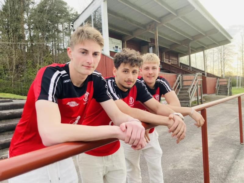 Tre unga män i röda fotbollströjor lutar sig mot ett räcke framför en fotbollsplan med läktare. Träd och en byggnad finns i bakgrunden. De tittar på kameran och verkar vara lagkamrater.