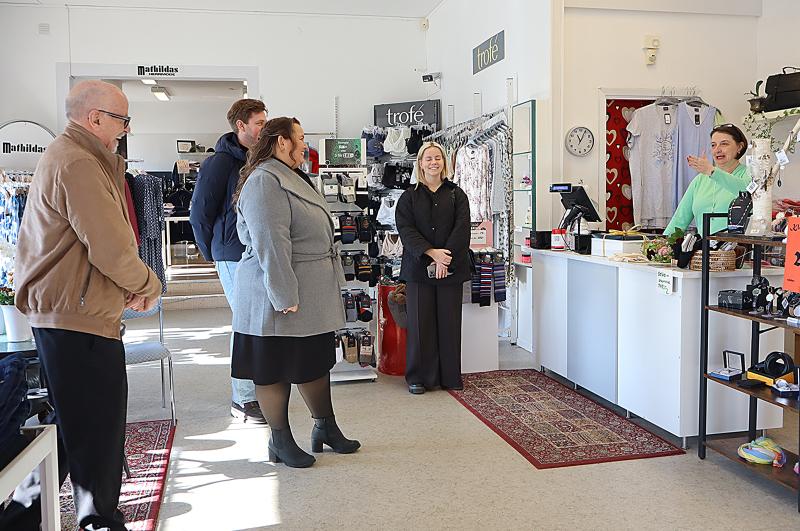 Fyra personer står vända mot en kassörska bakom en disk i en kläd- och accessoarbutik. Kassörskan talar och gestikulerar mot gruppen.