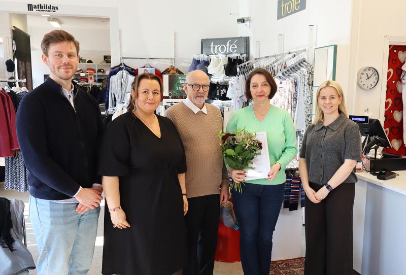 Fem personer står i en klädbutik; kvinnan i mitten håller i en blombukett och en tidning. Klädställ och en kassadisk syns i bakgrunden.