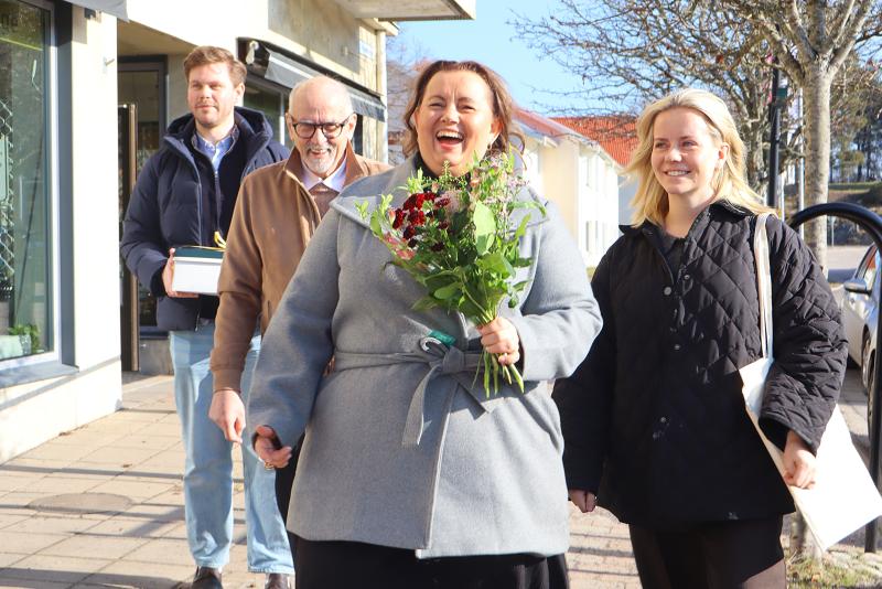 En kvinna med en blombukett går på en trottoar tillsammans med tre andra vuxna, varav en bär på en låda. De ser ut att le och promenera tillsammans en solig dag.