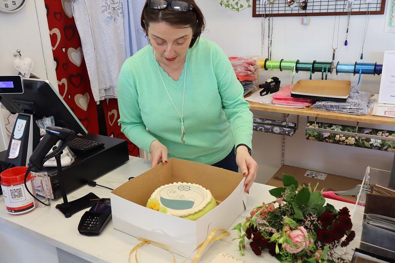En kvinna i grön tröja lägger en dekorerad tårta i en låda vid en butiksdisk, med blommor, en kassaapparat och förpackningsmaterial i närheten.