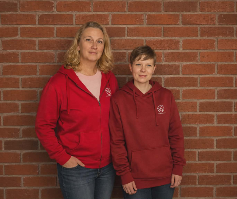 Två kvinnor står mot en röd tegelvägg, båda klädda i röda huvtröjor och blå jeans, vända mot kameran och poserar tillsammans.