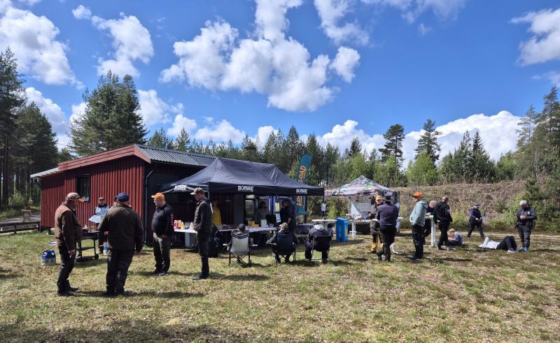 Människor samlas runt tält och bord som satts upp för ett utomhusevenemang nära en röd byggnad, omgiven av träd en solig dag med spridda moln.