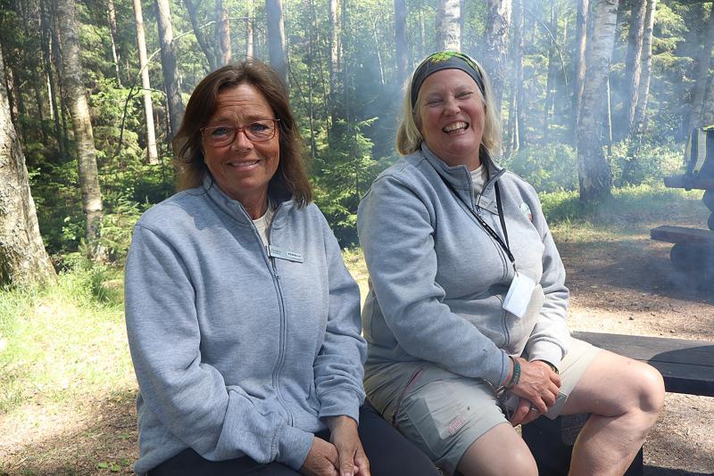 Två kvinnor med glasögon och grå jackor sitter på en bänk utomhus i ett skogsområde och ler mot kameran. I bakgrunden syns solljus och rök.