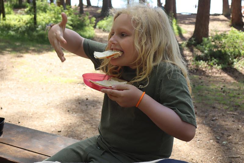 Ett barn med långt blont hår sitter utomhus på en träbänk i ett skogsområde och äter en tortilla, med en röd tallrik i handen.