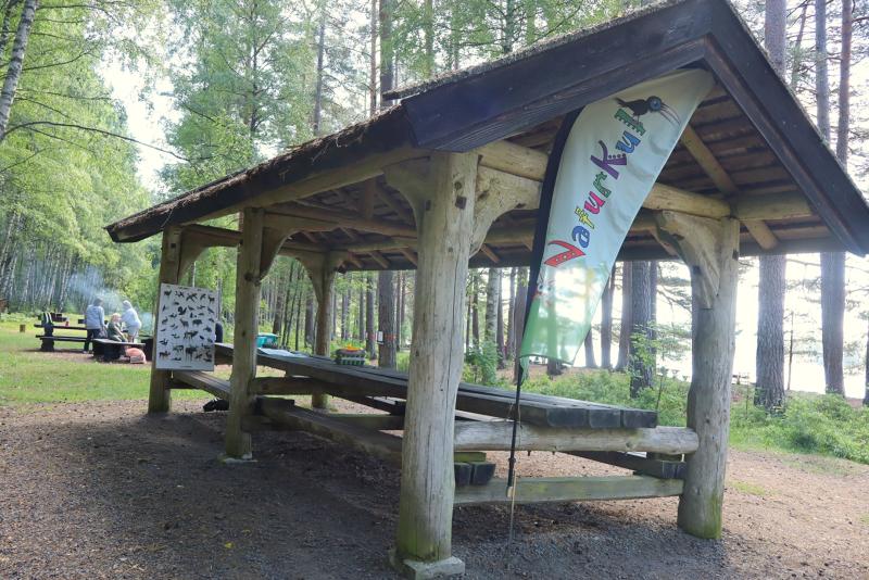 Ett picknicktak i trä med bänkar står i ett skogsområde nära en sjö, och där hänger en färgglad flagga samt affischer med fågelbestämning.
