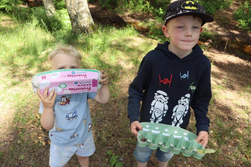 Två små pojkar står utomhus; den ene håller en rosa äggkartong framför ansiktet, och den andre håller en grön äggkartong med några små saker inuti. I bakgrunden syns träd och gräs.