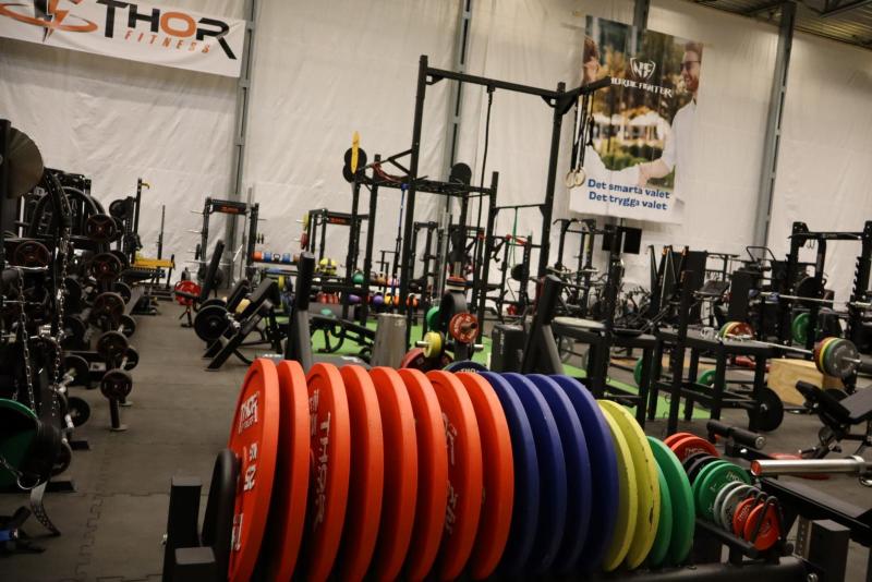 Ett gym med utrustning inklusive färgglada viktskivor, träningsmaskiner och skivstänger. En banderoll med människor som tränar hänger på väggen.