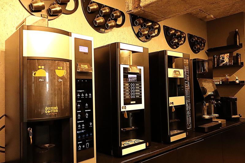 En rad kaffeautomater är placerade mot en vägg med monterade kaffekoppshållare och hyllor i ett café eller fikarum på ett kontor.