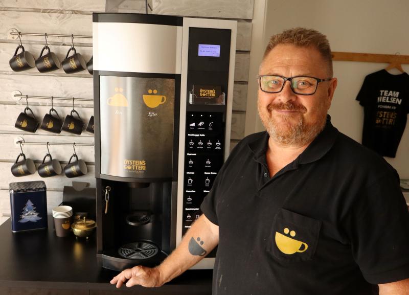 En man med glasögon och en svart skjorta med en kaffekoppslogotyp står bredvid en kaffemaskin, med muggar och andra kaffetillbehör i bakgrunden.