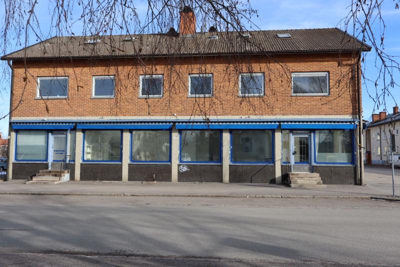 Tvåvånings tegelbyggnad med stängda skyltfönster och blå markiser, belägen på en lugn gata. Bladlösa trädgrenar ramar delvis in scenen.