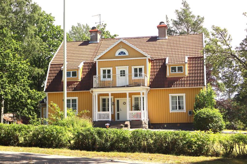 Ett tvåvåningshus i trä, gult till färgen, med rött tegeltak, vita lister och en veranda på framsidan, omgivet av grönska och träd.