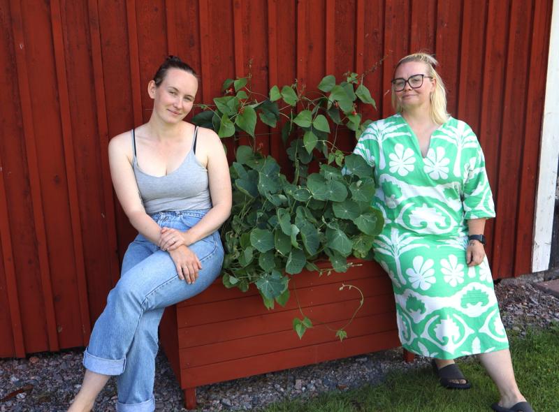 Två kvinnor sitter på vardera sidan om en planteringslåda med gröna bladväxter framför en röd trävägg.