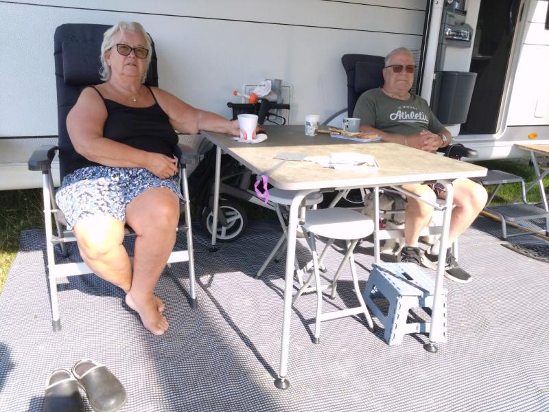 Två äldre vuxna sitter på stolar utanför en husbil, vid ett bord med drycker och tidningar, med en barnvagn och utomhusartiklar i närheten på en matta.