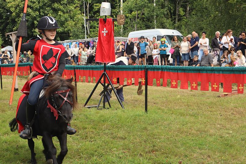 Ett barn i medeltida dräkt rider på en liten häst och håller i en lans under en tornerspelstävling, medan en folkmassa tittar på bakom en avspärrning prydd med röda baner.