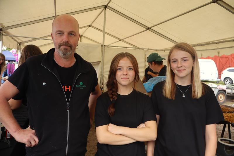 Tre personer i svarta tröjor står under ett tält vid ett utomhusevenemang, där två kvinnor står bredvid en man; i bakgrunden syns andra människor och tält.