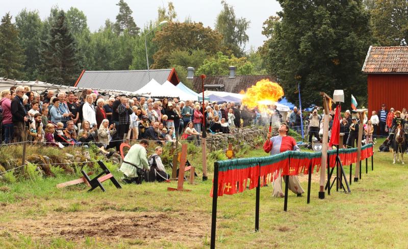 En medeltidsklädd artist blåser eld inför en stor publik på ett utomhusevenemang med gröna träd och röda byggnader i bakgrunden.