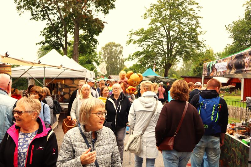 Människor går genom en utomhusmarknad med försäljningstält, träd och olika varor som visas upp. Vädret ser mulet ut.