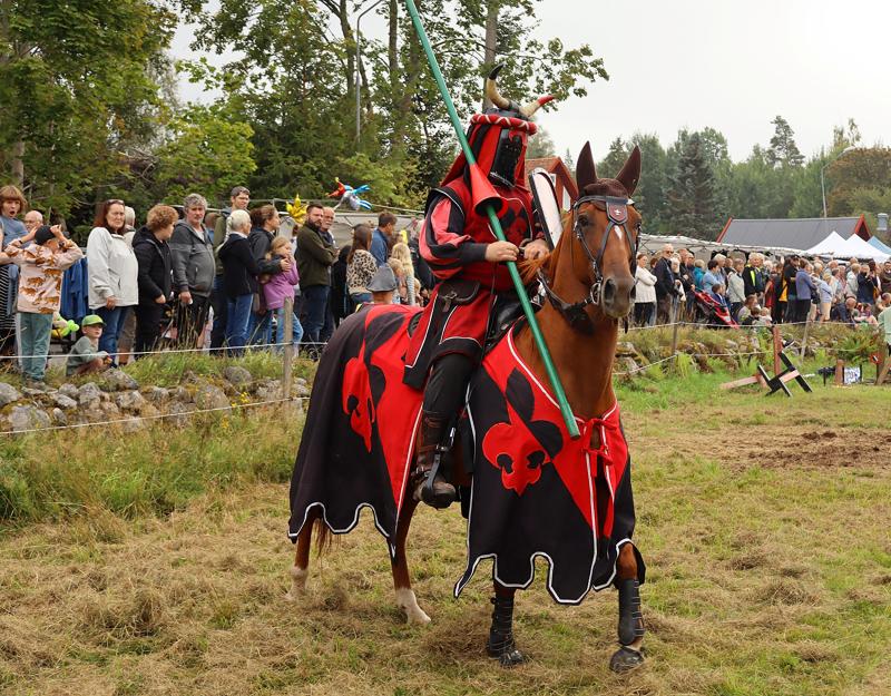 En person i röd och svart medeltida riddarrustning och hjälm rider på en häst klädd i matchande färger under ett offentligt utomhusevenemang med åskådare i bakgrunden.