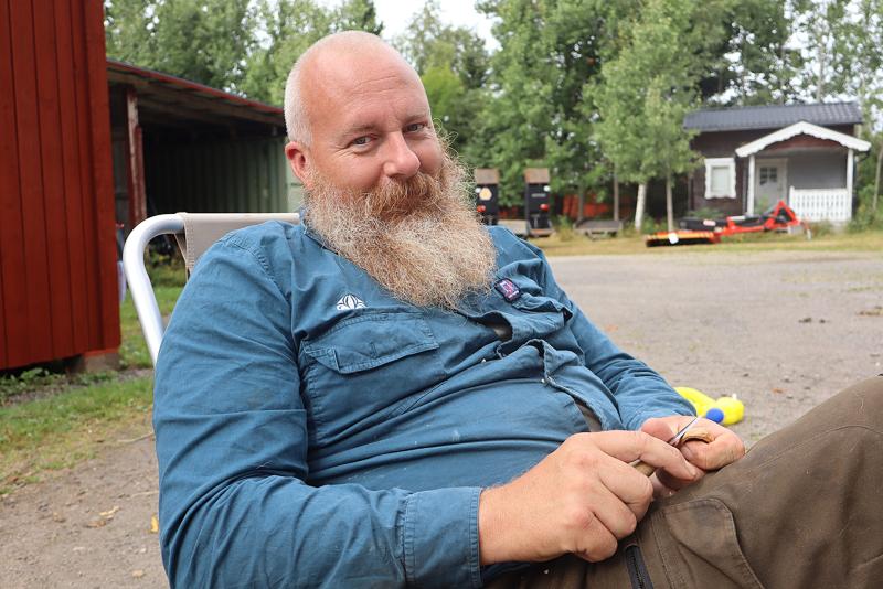 En skäggig man i blå skjorta sitter på en stol utomhus och håller ett föremål i händerna, med byggnader och träd i bakgrunden.