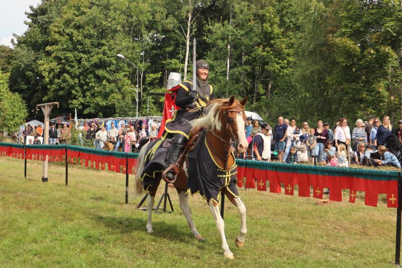 En person i medeltida rustning rider på en häst framför en folkmassa vid ett utomhusevenemang, med träd och röda baner i bakgrunden.