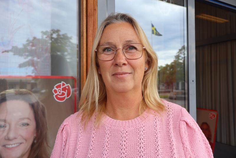 En kvinna med blont hår och glasögon har på sig en rosa tröja och står utomhus nära ett fönster, med affischer och en svensk flagga synliga i bakgrunden.